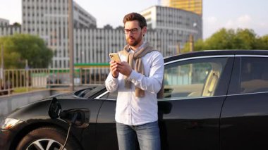 Telefonunu elektrikli aracının yanında tutan bir adam, şehir merkezinde şarj olurken gülümser..