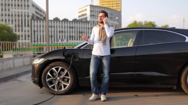 Elektrikli aracı şarj istasyonunda şarj olurken bir adam telefonuyla konuşur..