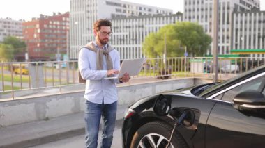 Bir adam elektrikli aracının yanında dizüstü bilgisayarını tutarken gülümsüyor..