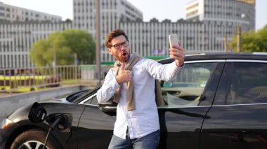 Bir adam, şarj istasyonunda şarj olurken elektrikli aracının yanında selfie çekerken gülümser..