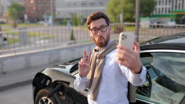 Bir adam, şarj istasyonunda şarj olurken elektrikli aracının yanında selfie çekerken gülümsüyor ve barış işareti yapıyor..