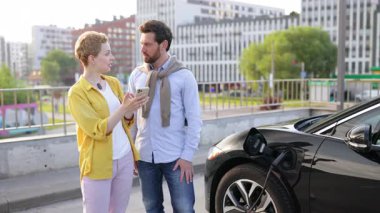 Gülümseyen bir çift şarj olurken elektrikli arabalarının yanında durur, muhtemelen akıllı telefon kullanarak süreci izlerler..