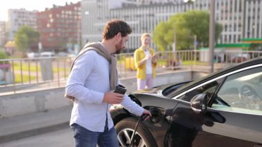 Bir erkek elektrikli araba şarj aletini takarken bir kadın arka planda onu izliyor..