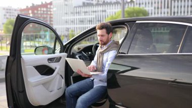 Gülümseyen bir adam, kapısı açık bir arabada otururken dizüstü bilgisayarında çalışır. Muhtemelen uzaktan çalışıyordur..