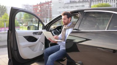 Siyah bir arabanın içindeki dizüstü bilgisayarda çalışırken gülümseyen bir adam kapıyı açık bırakıyor, muhtemelen bir şehir ortamında..