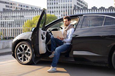 Bir iş adamı siyah bir arabada oturuyor ve modern bir binanın önünde kapısı açık bir şekilde dizüstü bilgisayarıyla çalışıyor..