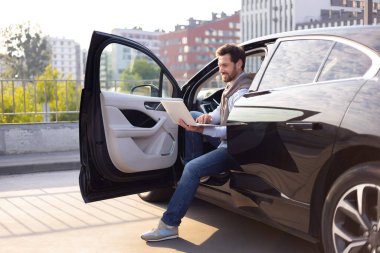 Siyah bir arabada oturan bir adam, dizüstü bilgisayarla çalışıyor, kapısı açık, muhtemelen şehir merkezinde..