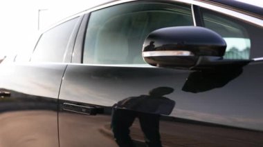 Bir adamın eli, parlak, siyah bir arabanın kapı koluna uzanır ve açmaya hazırdır..