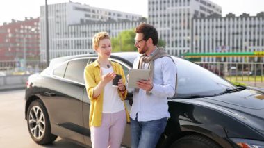 Gülümseyen bir çift şık, siyah bir arabanın yanında durur. Tablet tutan adam ve kahve fincanı tutan kadın..