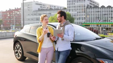 Gülümseyen bir çift siyah elektrikli bir arabanın yanında duruyor, başparmaklarını kaldırıyor, tablet ve kahve bardağıyla..