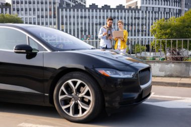 Bir erkek ve kadın, şehir merkezinde sade siyah bir elektrikli arabanın yanında dururken iş hakkında tartışıyorlar..