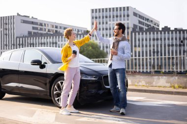 İki arkadaş, şehir ortamında parlak siyah bir elektrikli arabanın yanında beşlik çakarak kutluyor, muhtemelen başarılı bir sürüşten sonra..