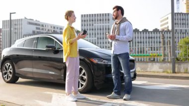 Bir kadın ve bir adam siyah elektrikli bir arabanın yanında el sıkışıyor, muhtemelen bir anlaşma ya da anlaşmadan sonra. Adamın elinde telefon, kadının elinde de kahve fincanı var..