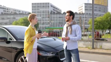 Siyah bir arabanın yanında bir erkek ve bir kadın, modern bir binanın önünde bir anı kutluyor..