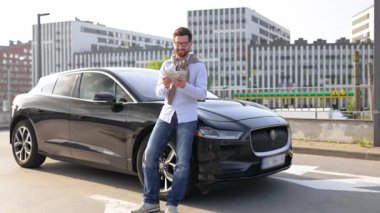 Gözlüklü bir adam, arka planında modern bir şehir manzarası olan gösterişli, siyah bir arabaya yaslanırken para sayar..