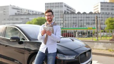 Elinde nakit para tutan gülümseyen bir adam şık, siyah bir arabanın yanında duruyor, muhtemelen bir alışverişi ya da finansal başarıyı kutluyor..