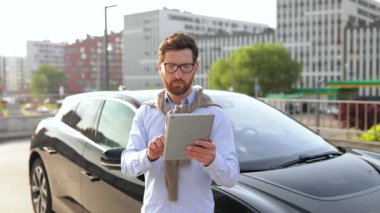 Neşeli bir adam şık, siyah bir arabanın yanında elinde bir tablet ve şehir ortamında bir başarıyı kutluyor..