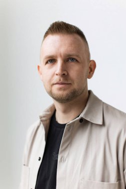 Kısa saçlı ve tarafsız bir yüz ifadesi olan bir adam bej bir gömlek içinde beyaz bir arka plana karşı poz verir..