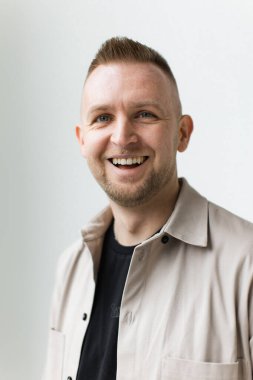 Mavi gözlü neşeli bir adam, bej bir gömlek giymiş, beyaz bir arka plana yaslanmış, geniş bir şekilde gülümsüyor..