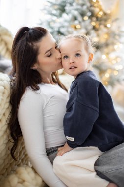 Bir anne küçük kızını yanağından öper, şefkatli bir an. Sahne sıcak ve sevgi dolu. Aile bağlarını göstermek için mükemmel..
