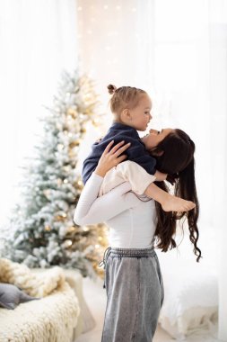 Bir anne, arka planda bir Noel ağacıyla hassas bir anı paylaşarak genç kızını kucağına alır..