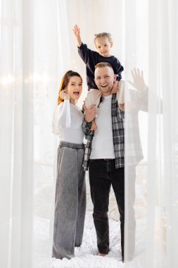 Üç kişilik neşeli bir aile birlikte poz veriyor, çocuğun babasının omuzlarında, yürek ısıtan bir sahne yaratıyor..