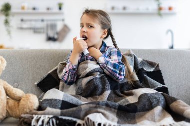 Battaniyeye sarılı genç bir kız, kanepede otururken astım spreyi kullanıyor, muhtemelen solunum hastalığından muzdarip..