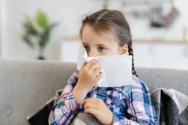 Örgülü genç bir kız kanepede oturuyor, burnuna mendil tutuyor, muhtemelen soğuk algınlığı ya da alerjiden muzdarip..