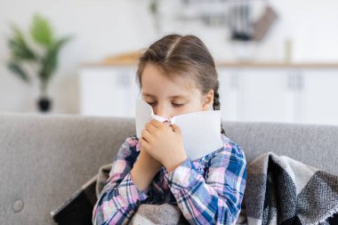 Battaniyeye sarılı genç bir kız, kanepede oturur, burnunu silmek için mendil kullanır, hasta olduğunu gösterir..