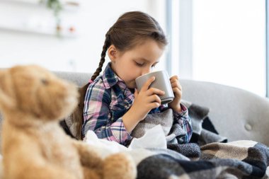 Muhtemelen hasta olan genç bir kız bir kanepede oturuyor ve bir kupadan su içiyor. Battaniyeye sarılmış, yanında oyuncak ayı var..