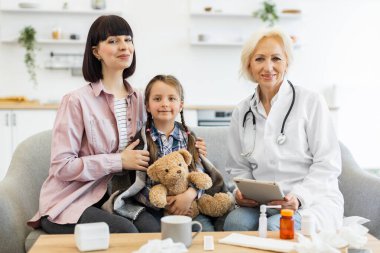 Bir anne ve kızı muayene için doktor muayenehanesindeler. Doktor gülümsüyor ve elinde bir tablet tutuyor..