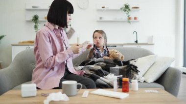 Endişeli bir anne telefonda konuşurken kızlarının sıcaklığını termometreyle ölçer, muhtemelen tıbbi tavsiye ister. Çocuk battaniyeye sarılmış..