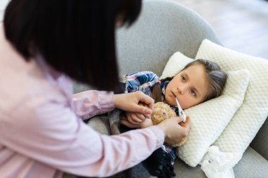 Endişeli bir anne hasta kızlarının sıcaklığını termometreyle ölçer. Kanepede dinlenir, bakım ve destek gösterir..