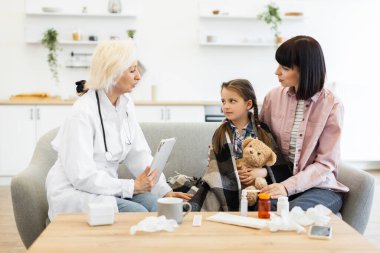 Bir bayan doktor, muhtemelen ev ziyareti sırasında bir kanepede genç bir kız ve annesine danışmanlık yapıyor. Çocuk hasta görünüyor, battaniyeye sarılmış ve oyuncak ayıyı tutuyor..
