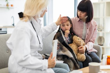 Endişeli bir doktor, annesi onu rahatlatırken genç kızların ateşini kontrol eder. Çocuk battaniyeye sarılmış ve oyuncak ayıyı tutuyor..