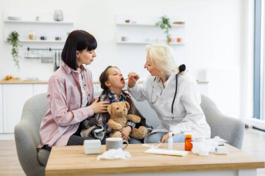 Endişeli bir anne, bir doktorun kızlarının boğazını muayene edişini izler, muhtemelen hastalık kontrolü yapar. Kız bir kanepede oyuncak ayıyla oturuyor..