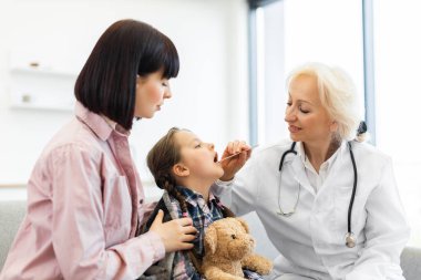 Bir kadın doktor, genç bir kızın boğazını incelemek için dil bastırıcı kullanır. Bu arada annesi de ona bakar..
