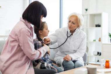 Kadın bir doktor, annesi yanında otururken genç bir kızın kalbini dinlemek için steteskop kullanıyor. Sahne bir doktorun ofisinde geçiyor..
