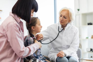 Kadın bir doktor, kontrol sırasında genç bir kızın kalbini dinlemek için steteskop kullanır. Annesi de destek olur..