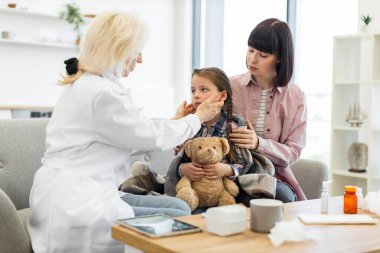 Annesi onu rahatlatırken bir doktor genç bir kızın boğazını kontrol ediyor. Çocuk battaniyeye sarılmış ve oyuncak ayıyı tutuyor..