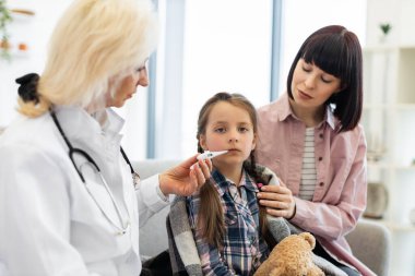 Annesi onu rahatlatırken bir doktor termometreyle genç bir kızın ateşini ölçer. Çocuk hasta görünüyor, battaniyeye sarılmış..