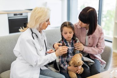 Endişeli bir doktor, annesi onu rahatlatırken genç bir kızın ateşini termometreyle ölçer. Sahne rahat bir evde geçiyor..
