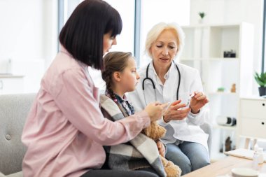 Beyaz önlüklü bir doktor, genç bir kıza ve annesine klinik ortamında tıbbi tavsiyelerde bulunan bir ilacı açıklar..