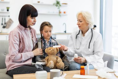 Bir doktor, oyuncak ayıyla battaniyeye sarılmış bir anneye ve hasta kızına ilaç gösteriyor..