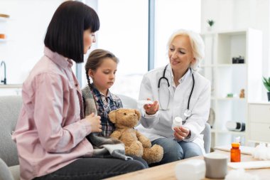 Bir doktor battaniyeye sarılmış ve muhtemelen bir klinikte oyuncak ayı tutan bir anneye ve kızına ilaç gösteriyor..