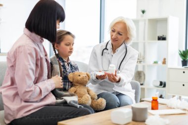 Bir doktor genç bir kıza ve annesine ilaç gösteriyor. Muhtemelen ilaçların dozajını ve kullanımını açıklıyor..