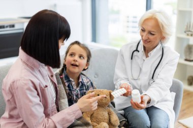 Bir kadın doktor genç bir kıza ve annesine ilaç gösteriyor. Muhtemelen bir hastalığın dozajını ve tedavi planını açıklıyor..