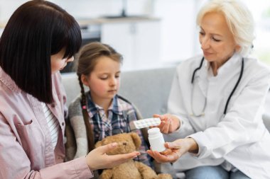 Bir doktor genç bir kıza ve annesine ilaç gösteriyor. Çocuk bir battaniyeye sarılmış, hasta görünüyor, doktor ise tedaviyi açıklıyor..