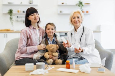 Bir doktor evde bir anneye ve hasta kızına danışmanlık yapıyor, çocuğa ilaç ve bakım öneriyor. Kız battaniyeye sarılmış..