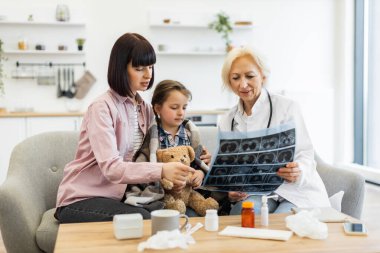 Bir doktor anne ve çocuğuyla birlikte röntgeni inceler, muhtemelen çocuk sağlığı ve teşhisini tıbbi bir ortamda tartışır..